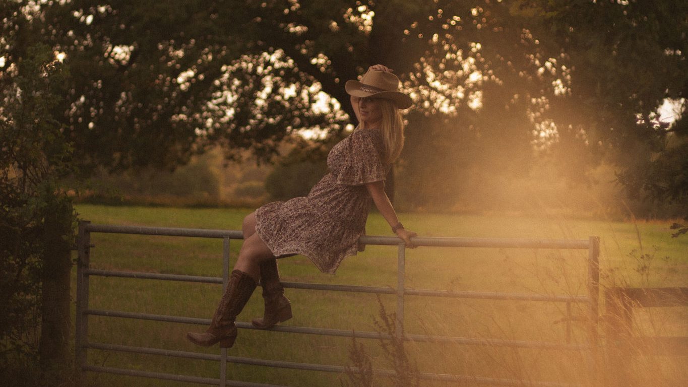 Cowgirl sitting on a fence in the countryside. Wearing Ariat Casanova boots and a cowboy hat.